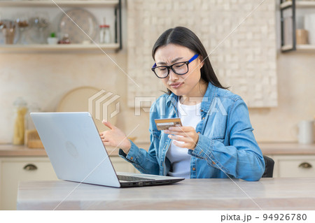 Upset and sad woman in depression trying to make online purchase in online store, Asian woman sitting at home in kitchen using laptop and bank credit card, unable to make payment online. Upset and sad woman in depression trying to make online purchase in online store, Asian woman sitting at home in kitchen using laptop and bank credit card, unable to make payment online. 94926780