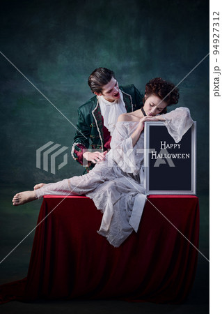 Portrait of young beautiful woman in white dress sitting on table with Halloween tablet and man in character of vampire biting her Portrait of young beautiful woman in white dress sitting on table with Halloween tablet and man in character of vampire biting her 94927312