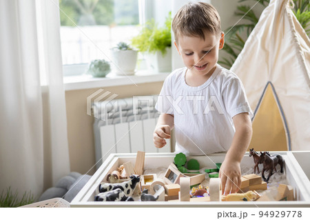 Smiling boy kid playing sensory box kinetic sand table with Montessori wooden materials Smiling boy kid playing sensory box kinetic sand table with Montessori wooden materials 94929778