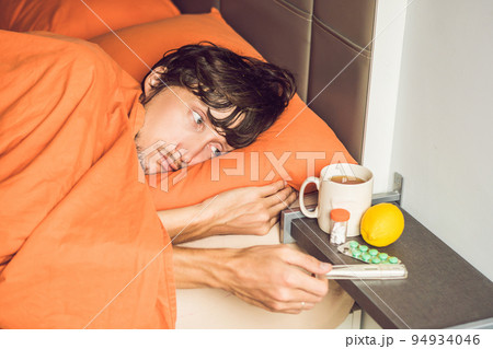 Man feeling cold, lying in the bed and drinks tea and measures temperature with a thermometer 94934046