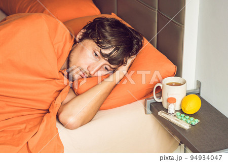 Man feeling cold, lying in the bed and drinks tea and measures temperature with a thermometer 94934047