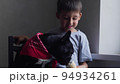 boy child sits next to a black French bulldog and feeds ice cream in a cup. sitting at home next to the window of the house 94934261