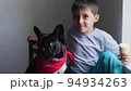 boy child sits next to a black French bulldog and feeds ice cream in a cup. sitting at home next to the window of the house 94934263