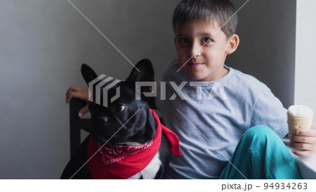 boy child sits next to a black French bulldog and feeds ice cream in a cup. sitting at home next to the window of the house boy child sits next to a black French bulldog and feeds ice cream in a cup. sitting at home next to the window of the house 94934263