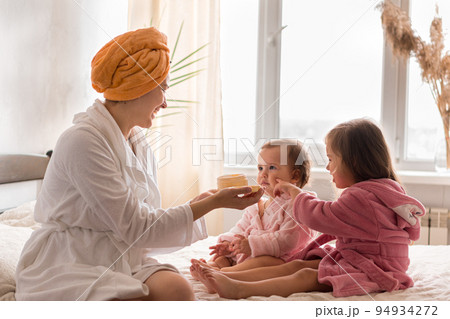 Young mother two daughters in curlers bathrobes. happy smiling family skin care concept. Mom teaches little kids child to do makeup smear cream on face sittting in orange bathrobes on bed in bedroom 94934272