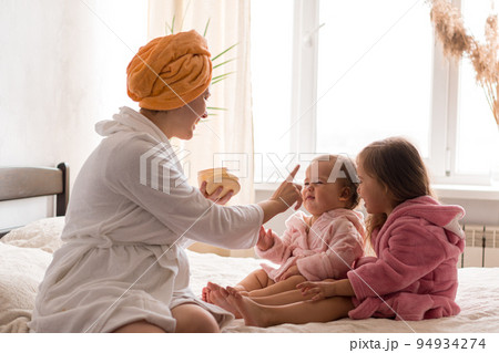 Young mother two daughters in curlers bathrobes. happy smiling family skin care concept. Mom teaches little kids child to do makeup smear cream on face sittting in orange bathrobes on bed in bedroom 94934274