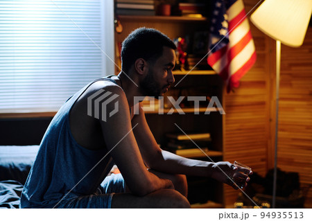 Young depressed man with glass of whisky sitting on bed in bedroom 94935513