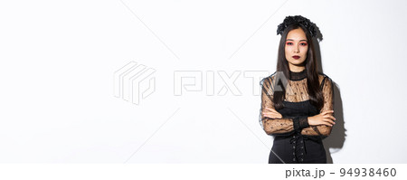 Image of beautiful asian woman in black lace dress and wreath looking serious. Girl dressed-up for halloween party as evil witch, standing over white background 94938460