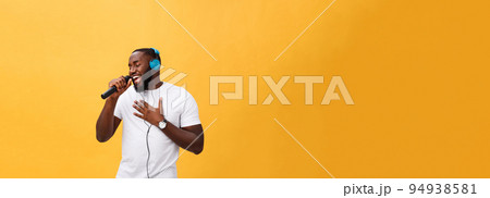 Portrait of cheerful positive chic handsome african man holding microphone and having headphones on head listening music singing song enjoying weekend vacation isolated on yellow background 94938581