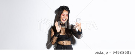Portrait of beautiful and stylish asian woman in gothic lace dress saying hi, waving hand at smartphone camera during video call, standing over white background Portrait of beautiful and stylish asian woman in gothic lace dress saying hi, waving hand at smartphone camera during video call, standing over white background 94938685