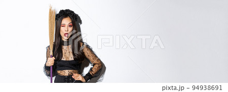Beautiful asian woman celebrating halloween in witch costume, holding broom and showing tongue, winking silly at camera, standing over white background 94938691
