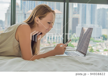 Young woman is working on a laptop in his bed on a background of a panoramic window overlooking the skyscrapers. Freelancer, remote work, work from home Young woman is working on a laptop in his bed on a background of a panoramic window overlooking the skyscrapers. Freelancer, remote work, work from home 94938822