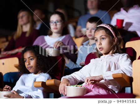 Girls watching a movie and eating popcorn in cinema Girls watching a movie and eating popcorn in cinema 94943581