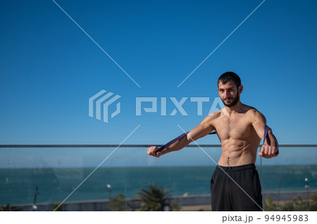 Shirtless man doing exercise with rubber bands outdoors.  94945983