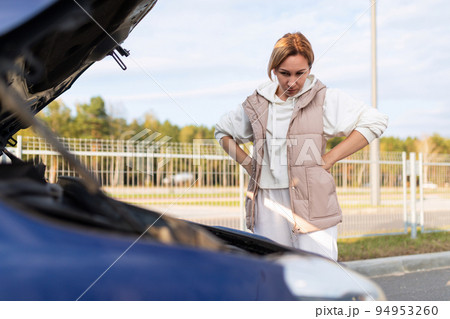 a young woman at the open hood of a car is trying to figure out the malfunction of the car, car a young woman at the open hood of a car is trying to figure out the malfunction of the car, car 94953260