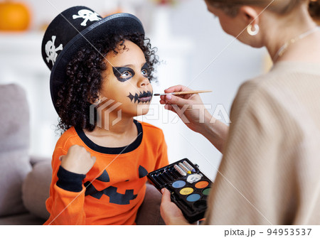 Festive makeup for Halloween. Woman doing pumpkin make-up for ethnic curly boy in costume while preparing holiday 94953537