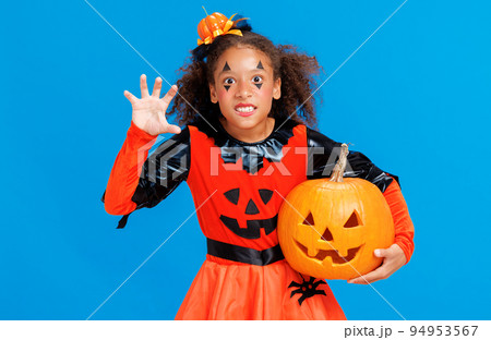 Happy Halloween ethnic girl in a pumpkin costume with jack-o-lantern scary gestures on blue background Happy Halloween ethnic girl in a pumpkin costume with jack-o-lantern scary gestures on blue background 94953567
