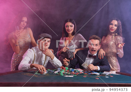 Friends playing poker at casino, at table with stacks of chips, money, cards on it. Celebrating win, smiling. Black, smoke background. Close-up. Friends playing poker at casino, at table with stacks of chips, money, cards on it. Celebrating win, smiling. Black, smoke background. Close-up. 94954373