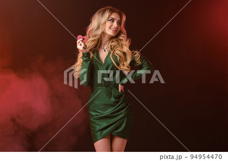 Blonde girl in green stylish dress and jewelry. Smiling, showing two red chips, hand on waist, posing on colorful smoky background. Close-up 94954470