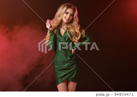 Gorgeous blonde lady in green stylish dress and jewelry. Smiling, showing two red chips, hand on waist, posing on colorful smoky background. Close-up 94954471