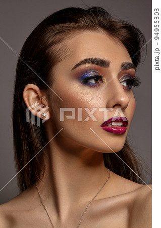 Brunette naked woman in jewelry posing on gray studio background. Luxury makeup. Colorful eyeshadow, long eyelashes, glossy red lips. Close up 94955303