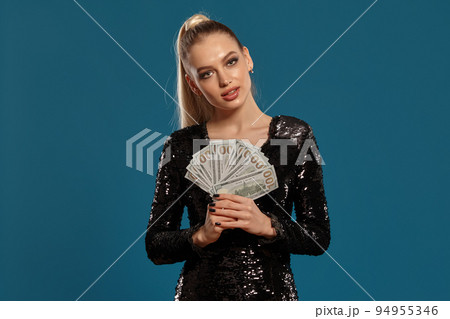 Blonde lady with ponytail, in black sequin dress. Showing fan of hundred dollar bills, posing on blue background. Gambling, poker, casino. Close-up Blonde lady with ponytail, in black sequin dress. Showing fan of hundred dollar bills, posing on blue background. Gambling, poker, casino. Close-up 94955346