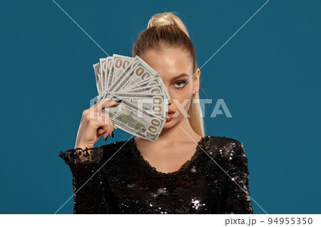 Blonde woman with ponytail, in black sequin dress. Showing fan of hundred dollar bills, posing on blue background. Gambling, poker, casino. Close-up 94955350