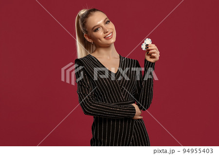 Blonde lady in jewelry and black dress. She smiling, hugging her own waist, showing two white chips, posing on red background. Poker, casino. Close-up 94955403