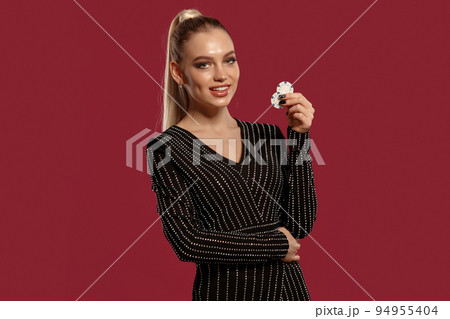 Blonde model in jewelry and black dress. Smiling, hugging her own waist, holding two white chips, posing on red background. Poker, casino. Close-up 94955404