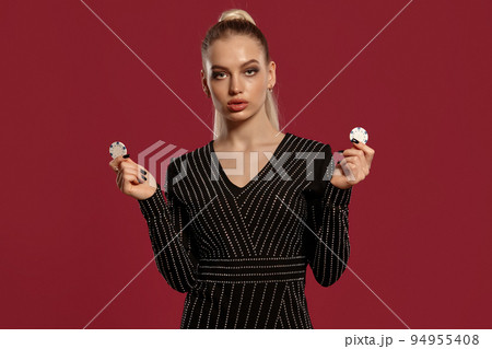 Blonde woman with ponytail, in black dress in rhinestones. Showing two white chips, posing on red background. Gambling, poker, casino. Close-up 94955408