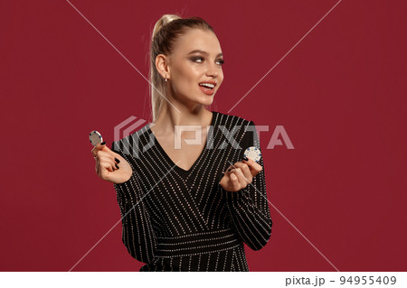 Blonde female with ponytail, in black dress in rhinestones. She smiling, showing two white chips, posing on red background. Poker, casino. Close-up 94955409
