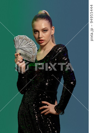 Blonde girl in jewelry and black sequin dress. She is showing some cash, posing on colorful background. Poker, casino. Close-up 94955444