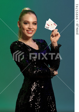 Blonde girl in jewelry and black sequin dress. She smiling, showing two playing cards, posing on colorful background. Poker, casino. Close-up Blonde girl in jewelry and black sequin dress. She smiling, showing two playing cards, posing on colorful background. Poker, casino. Close-up 94955741
