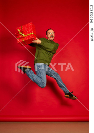 Portrait of young cheerful man jumping in joy with present box isolated over red studio background. Super excited 94958812