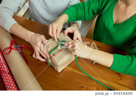 Top view image of man and woman wrapping presents together at home, using wrapping paper and ribbons. Making bow with string Top view image of man and woman wrapping presents together at home, using wrapping paper and ribbons. Making bow with string 94958890