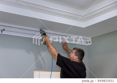 There is a professional handyman who is nailing wooden cornices on the curtain wall over the window of a newly constructed home. There is a professional handyman who is nailing wooden cornices on the curtain wall over the window of a newly constructed home. 94965931