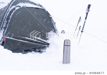 Tourist tent in the snow. Thermos and trekking poles in the snow. Tourist equipment. Snowfall on a hike. Hike in winter. 94968677