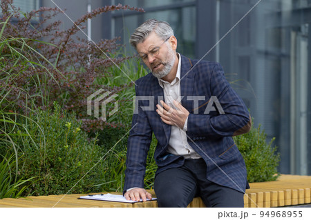 Heartache of mature man, gray haired businessman holding hands on rough, heart attack pain, boss outside modern office building on bench, sick and overworked. 94968955