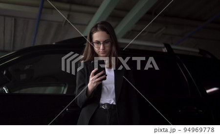 A young businesswoman is standing in the parking lot by the car with a phone. A girl with glasses and a jacket is texting on a smartphone. A young businesswoman is standing in the parking lot by the car with a phone. A girl with glasses and a jacket is texting on a smartphone. 94969778