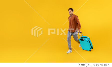 Happy Tourist Guy Walking With Blue Suitcase Over Yellow Background Happy Tourist Guy Walking With Blue Suitcase Over Yellow Background 94970307