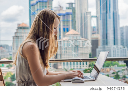 Young woman is working on a laptop on her balcony overlooking the skyscrapers. Freelancer, remote work, work from home Young woman is working on a laptop on her balcony overlooking the skyscrapers. Freelancer, remote work, work from home 94974326