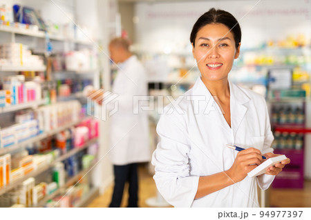 Positive female pharmacist writes in a notebook in the trading floor of pharmacy Positive female pharmacist writes in a notebook in the trading floor of pharmacy 94977397