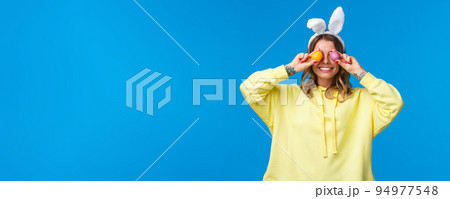 Holidays, traditions and celebration concept. Happy carefree smiling caucasian woman celebrating Easter, holding two painted eggs on eyes and grinning, wear cute rabbit ears, blue background Holidays, traditions and celebration concept. Happy carefree smiling caucasian woman celebrating Easter, holding two painted eggs on eyes and grinning, wear cute rabbit ears, blue background 94977548