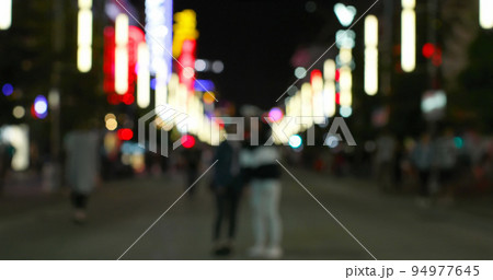 Image of defocussed shop and street lights, with people walking on a city road at night 94977645