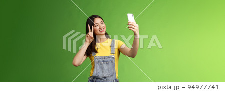 Tender cute asian brunette female, hold smartphone, posing selfie, look telephone screen, photographing, show victory peace sign, joyfully chatting friend abroad, stand green background 94977741