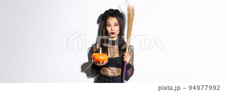 Image of sassy witch in gothic lace dress, holding broom and pumpkin, looking at upper left corner with halloween banner, white background Image of sassy witch in gothic lace dress, holding broom and pumpkin, looking at upper left corner with halloween banner, white background 94977992
