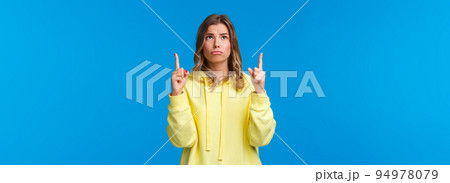 Jealous and gloomy cute blond female student feel regret not having something she desires, sobbing frowning sad as looking pointing up, standing over blue background 94978079
