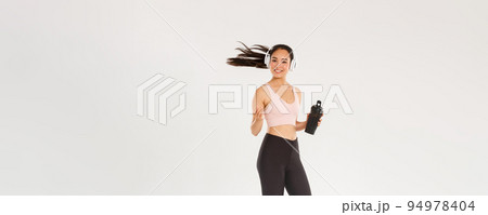 Full length of smiling attractive asian female athelte, girl doing fitness workout in gym, sportswoman or runner drinking water from bottle, listening music in wireless headphones, tossing hair Full length of smiling attractive asian female athelte, girl doing fitness workout in gym, sportswoman or runner drinking water from bottle, listening music in wireless headphones, tossing hair 94978404