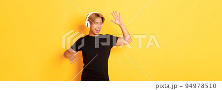 Happy attractive asian guy with blond hair, singing along and dancing as listening to music in wireless headphones, standing yellow background 94978510