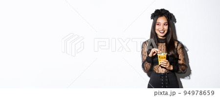 Smiling cute asian woman celebrating halloween, holding sweets and grinning happy, trick or treating in witch costume 94978659